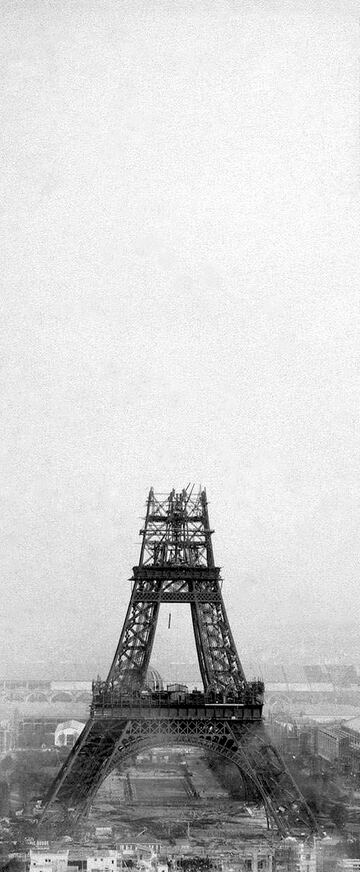 Photographie ancienne de la tour Eiffel en construction : les quatre piliers sont réunis au-dessus du premier étage, la partie haute n'est pas encore montée, des grues sont visibles au sommet.
