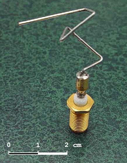 Une petite antenne en fil métallique plié en zigzag irrégulier, fixée sur un connecteur doré, avec une échelle de 2 cm.