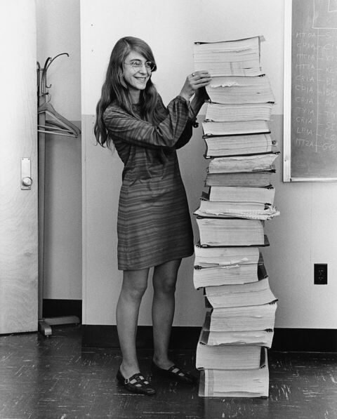 Photographie en noir et blanc d'une jeune femme debout à côté d'une pile de classeurs de listings aussi haute qu'elle.
