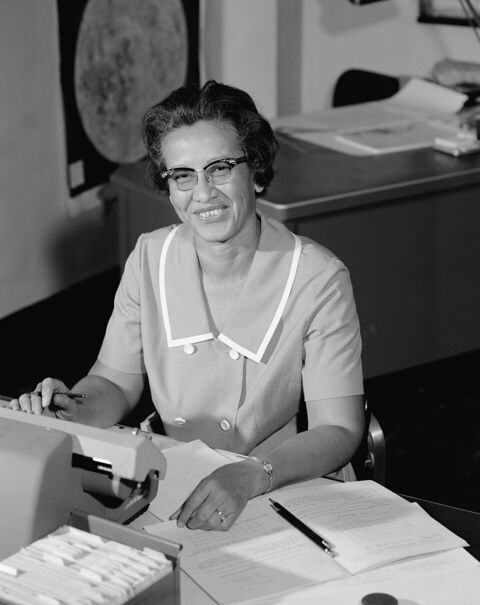 Photographie en noir et blanc d'une femme assise à un bureau, souriante, devant une machine à calculer et des documents.