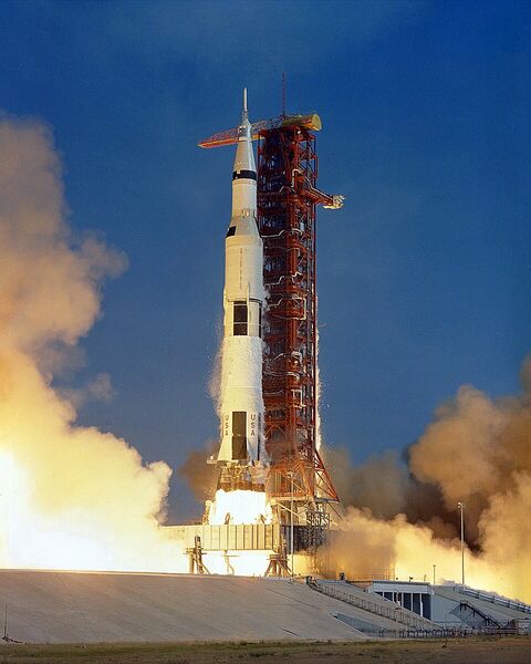 Photographie du décollage d'une fusée Saturn V blanche et noire, très haute, au milieu d'un nuage de fumée et de flammes, à côté de la tour de lancement.