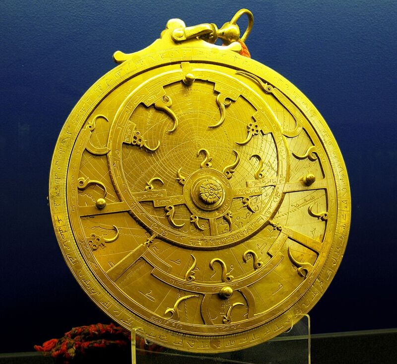 Astrolabe persan en laiton : sur le fond gris gravé de cercles, une araignée ajourée dorée porte de petites pointes recourbées, et une règle tourne autour du centre.