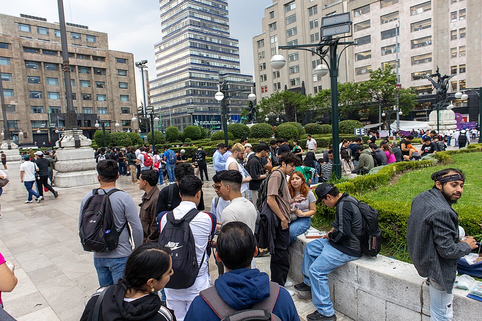 Sur une place de Mexico, des personnes assises et debout comparent et échangent des vignettes autour d'albums ouverts.