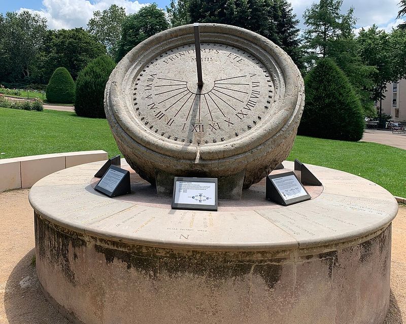 Cadran solaire de pierre en forme d'anneau incliné posé sur un socle circulaire dans un parc ; une tige traverse le centre de l'anneau gradué.