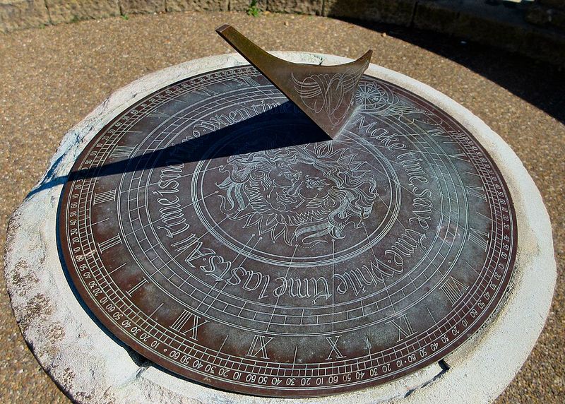 Cadran solaire horizontal en bronze posé sur un socle de pierre : une plaque triangulaire dressée au-dessus du disque gradué projette une ombre nette sur les chiffres.