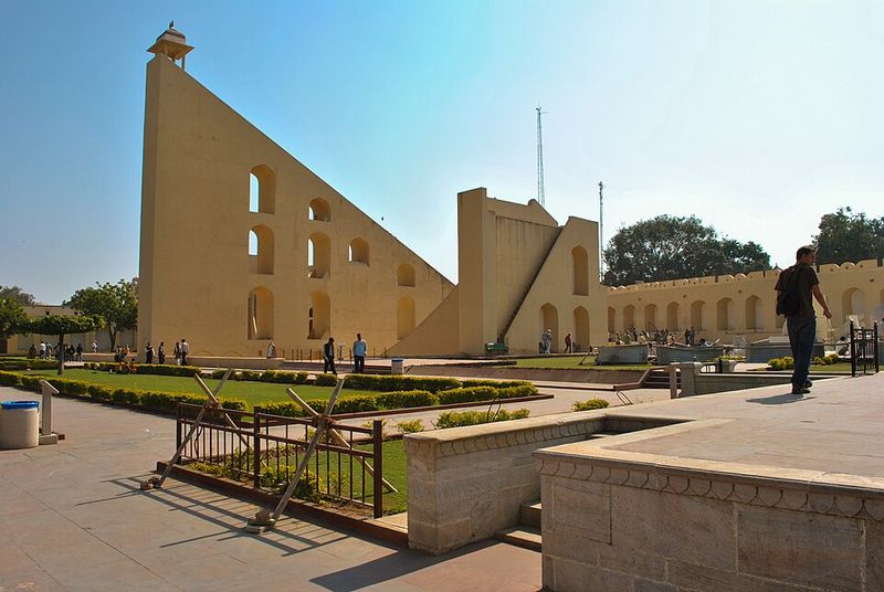 Gigantesque construction de pierre ocre en forme de triangle rectangle, avec un escalier montant le long de l'hypoténuse, entourée de deux quarts de cercle maçonnés, dans un jardin ensoleillé.