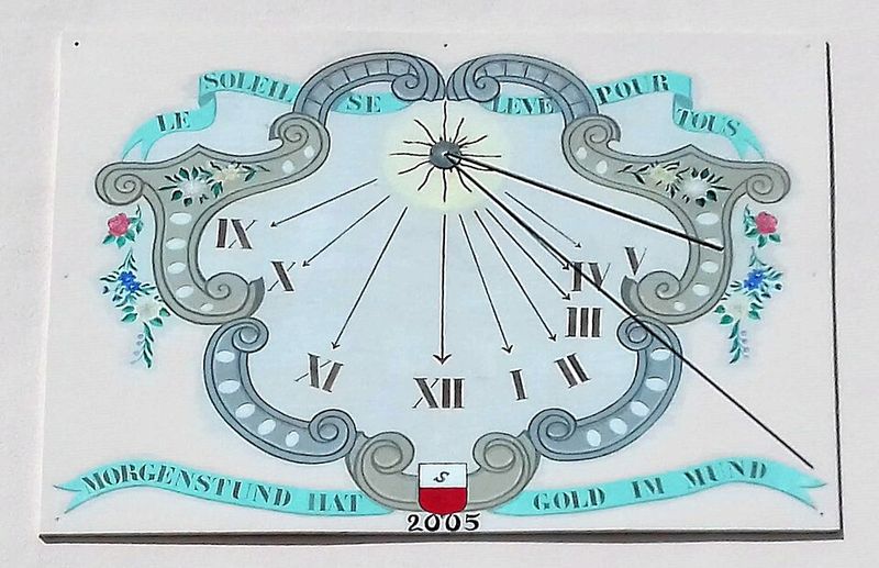 Cadran solaire peint sur un mur blanc : un cartouche décoré entoure des lignes horaires qui rayonnent depuis le pied d'une tige métallique, avec des chiffres romains de six à six.