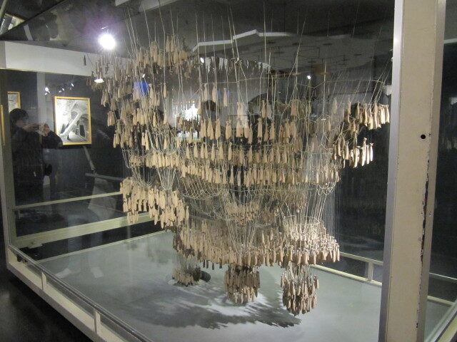 Dans une vitrine de musée, un enchevêtrement de fils suspendus au plafond, lestés de nombreux petits sacs, forme une structure à l'envers qui évoque une église.