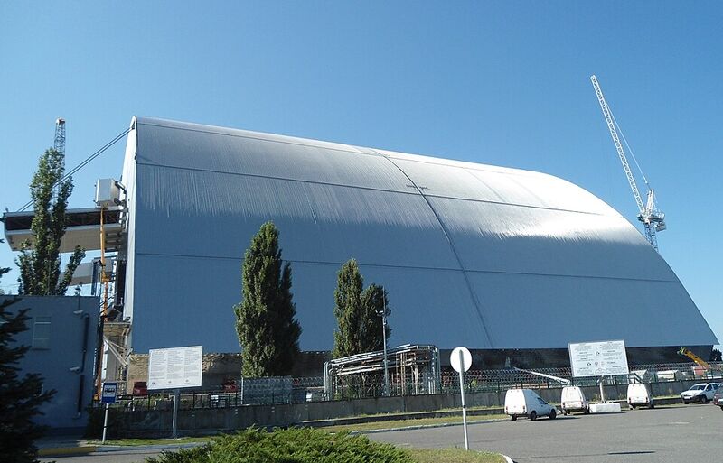 Photographie d'une immense arche métallique grise, de forme arrondie, posée sur le site d'une centrale nucléaire, avec une grue à droite.