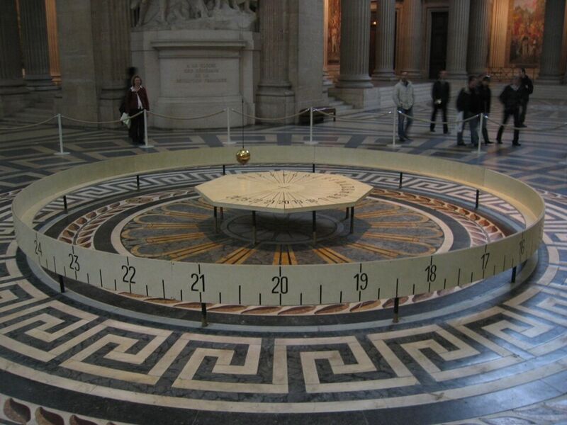 Photographie de l'intérieur du Panthéon : une boule dorée suspendue au-dessus d'une table circulaire, entourée d'une barrière graduée en heures.