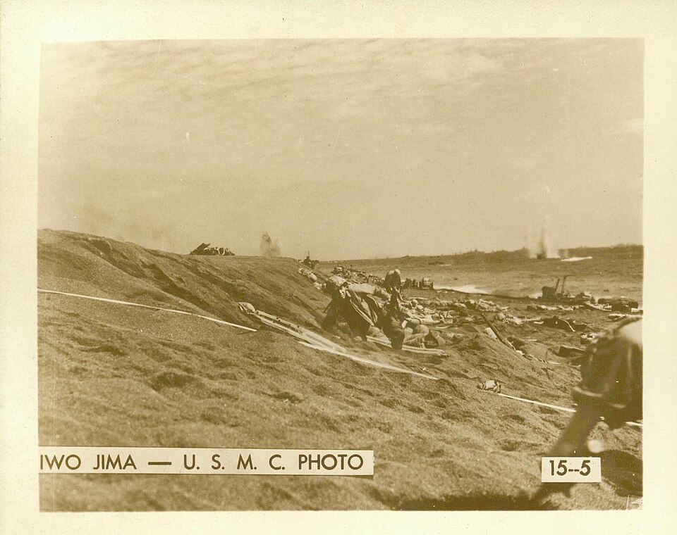 Photographie en noir et blanc : des soldats américains débarquent sur une plage de sable volcanique, avec des engins de débarquement et de la fumée à l'arrière-plan.
