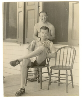 Photographie ancienne en noir et blanc : deux hommes sous un porche en bois, l'un debout, l'autre assis sur une chaise, les jambes croisées.