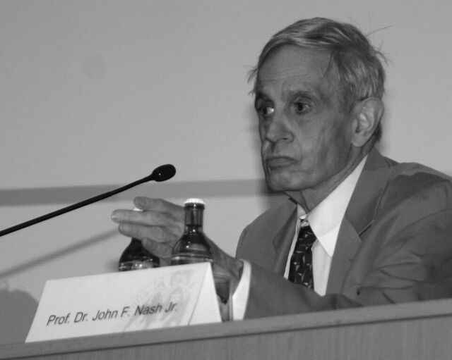 Photographie en noir et blanc d'un homme âgé en costume, assis à une table de conférence derrière un micro et un carton portant son nom.