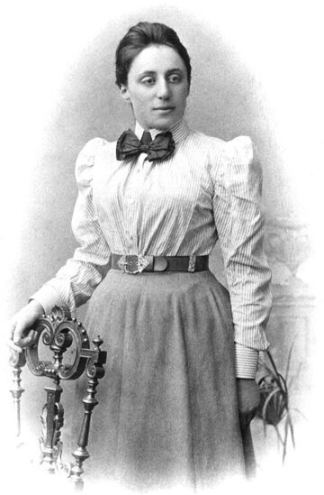Photographie ancienne en noir et blanc d'une jeune femme en robe sombre, les cheveux relevés, debout, de trois quarts.