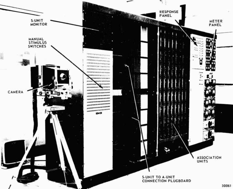Photographie en noir et blanc d'une grande armoire électronique avec des panneaux de câblage, et un appareil photo sur trépied devant.