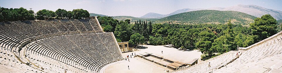 Vue panoramique du grand théâtre antique d'Épidaure : un hémicycle de gradins de pierre adossé à une colline boisée, autour d'une scène circulaire.