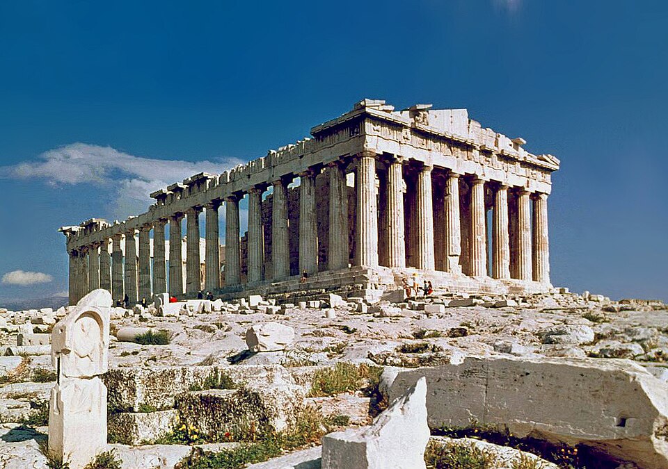Le Parthénon sur l'Acropole d'Athènes vu de trois quarts : une rangée de colonnes doriques sous un fronton en partie détruit, sur fond de ciel bleu.