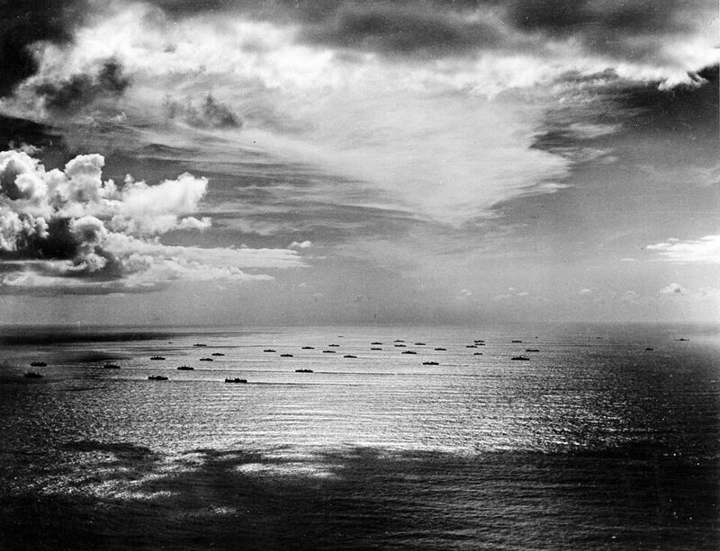 Photographie aérienne en noir et blanc : une quarantaine de navires dispersés en rangs sur l'océan, sous un ciel nuageux.