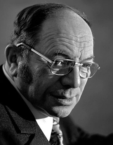 Photographie en noir et blanc d'un homme âgé à lunettes, en costume, souriant légèrement.