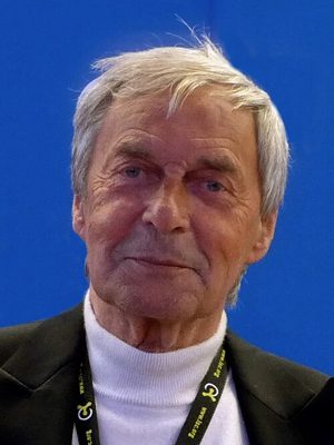 Portrait d'Ernő Rubik, homme âgé aux cheveux gris, en veste sombre, souriant légèrement.