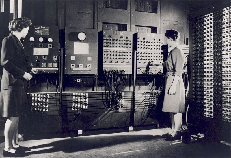Photographie en noir et blanc : deux femmes debout devant de grandes armoires de l'ENIAC couvertes de câbles, de prises et de boutons.