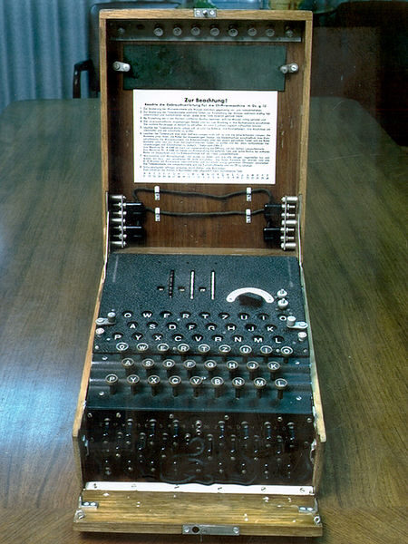 Photographie d'une machine Enigma dans sa caisse en bois ouverte : un clavier de lettres, au-dessus un tableau de lampes, trois rotors en haut, et à l'avant un tableau de prises reliées par des câbles.
