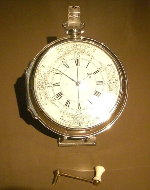 Photographie d'une grosse montre de poche en argent, au cadran blanc orné, exposée dans une vitrine sombre, avec sa clé de remontage.
