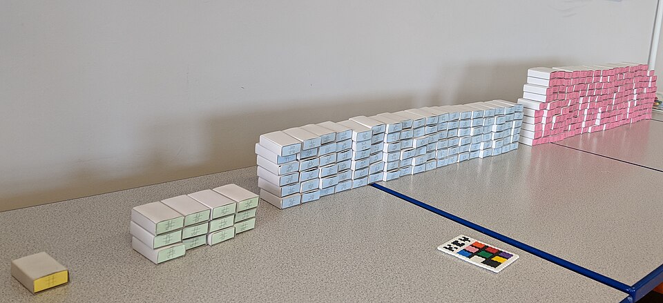 Une reconstitution de MENACE posée sur une table : une boîte jaune seule, un petit tas de boîtes vertes, un long mur de boîtes bleues et un mur de boîtes roses, chacune marquée d'une grille de tic-tac-toe.